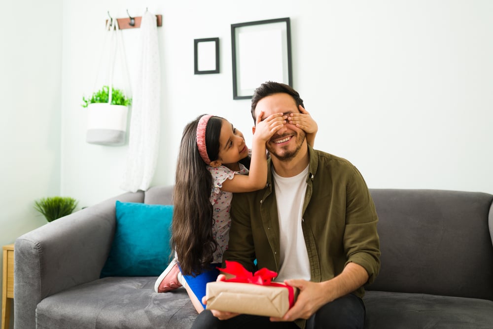 A happy dad sits on a couch with a gift while his daughter puts her hands over his eyes before he opens his present