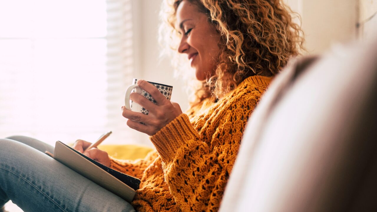 Happy woman holds tea while writing in journal and sitting on couch.