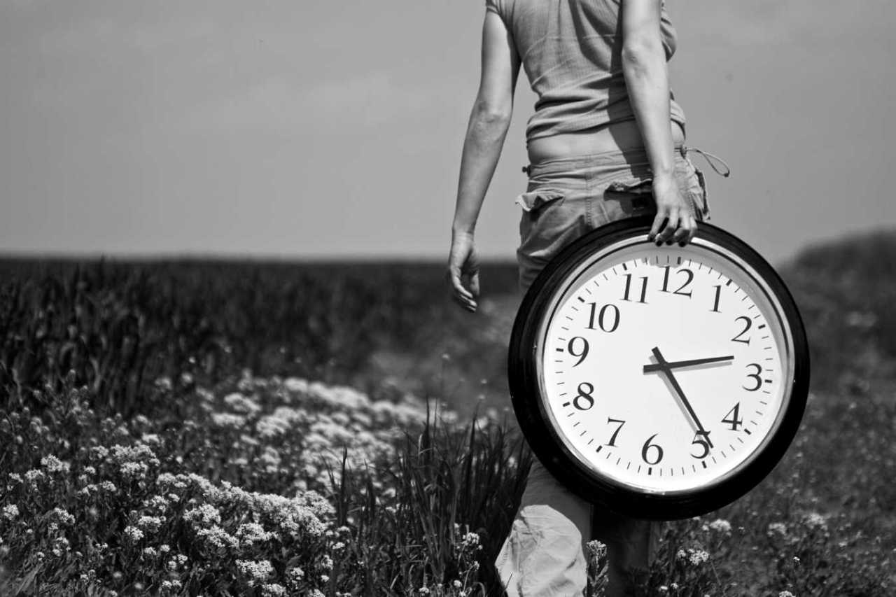 Young woman carrying a big clock on a field.