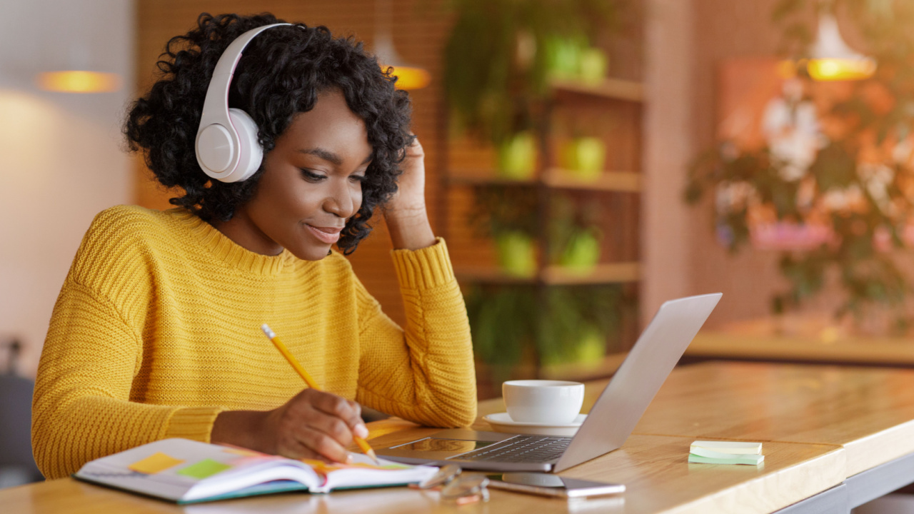 Smiling black girl with wireless headset studying online, using laptop at cafe.