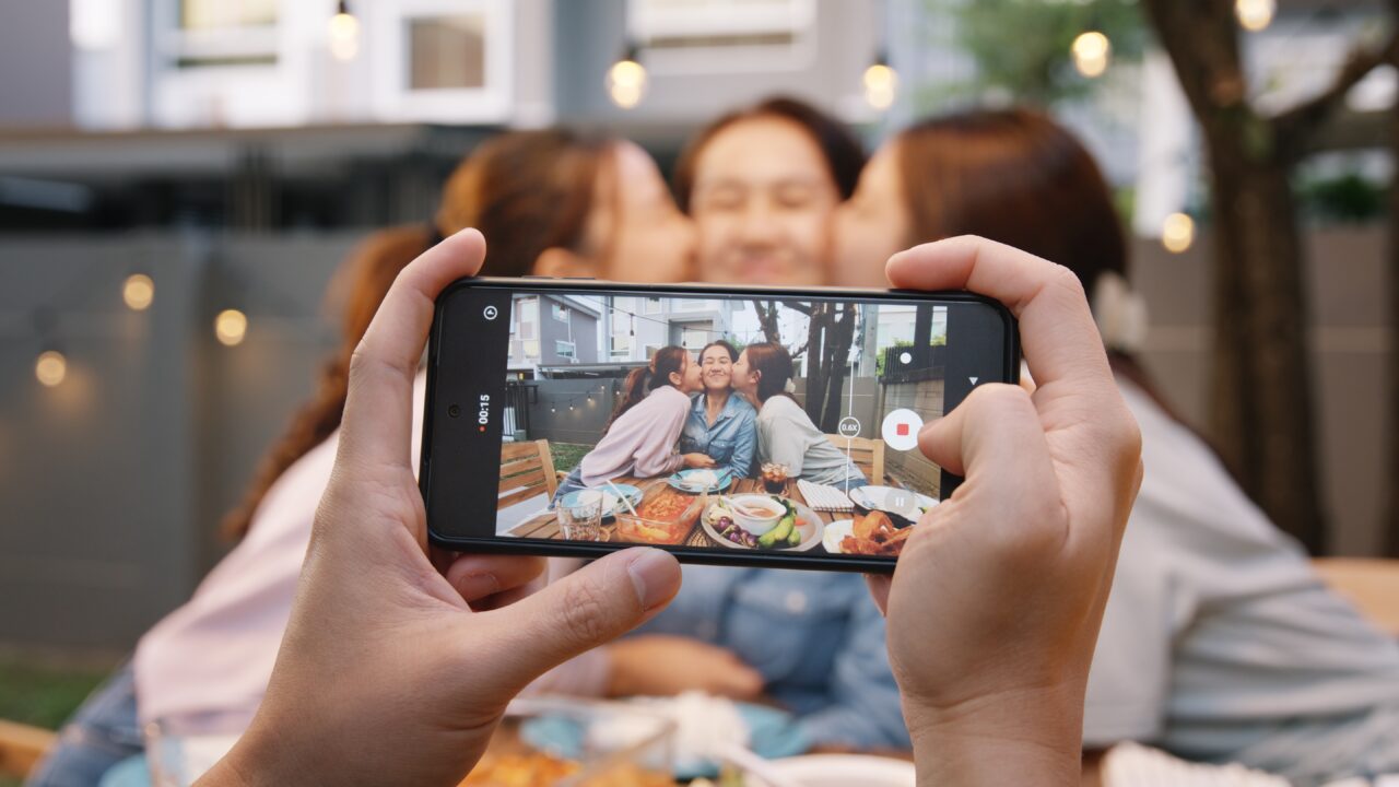 Hands hold a phone taking picture of three happy people at a table.