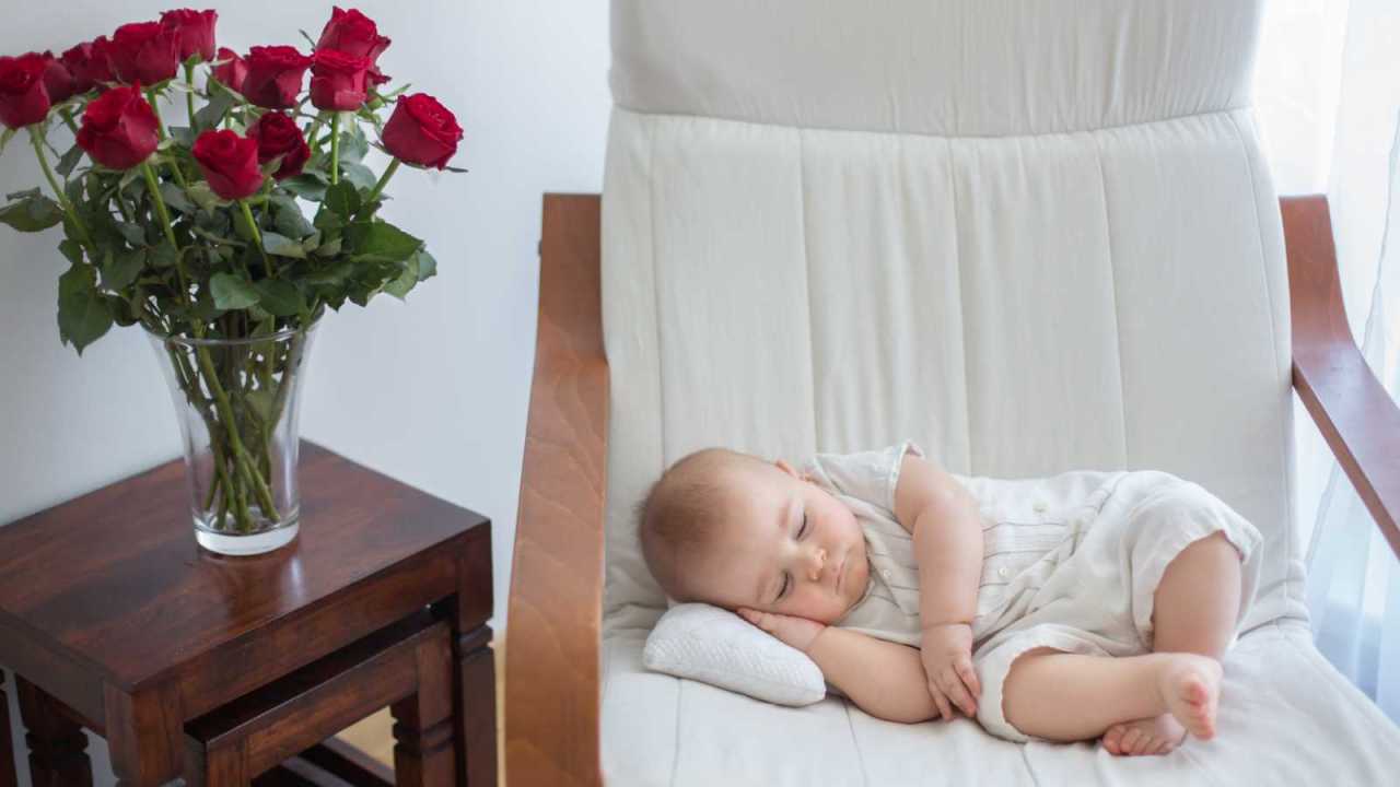 Baby asleep on chair next to table with vase of roses.