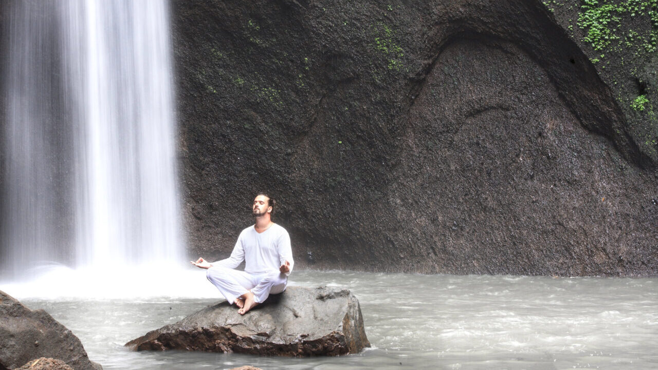 Man sitting on rock near a waterfall meditating.