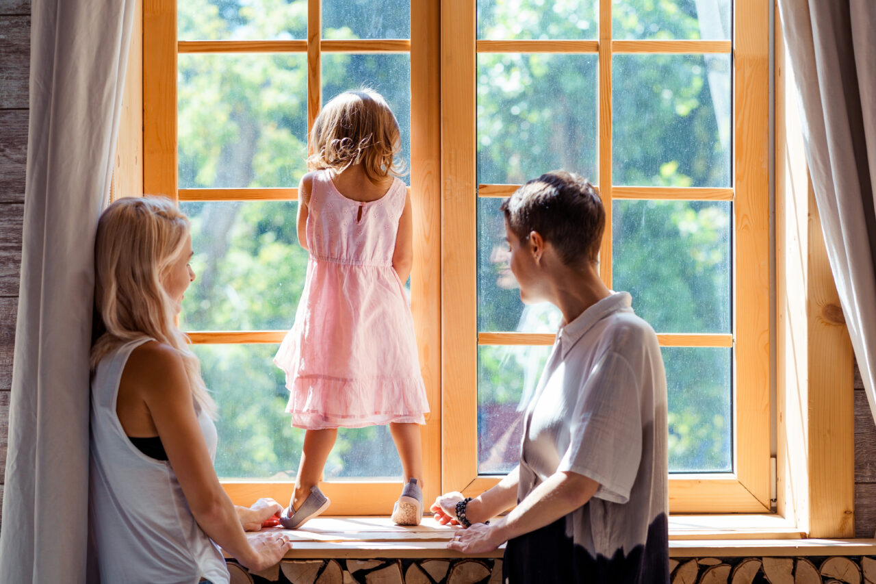 Lesbian mothers and their daughter standing near the window.