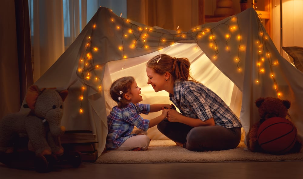 Mom and child bonding by playing in atent together. They are smiling and there are glowing fairy lights