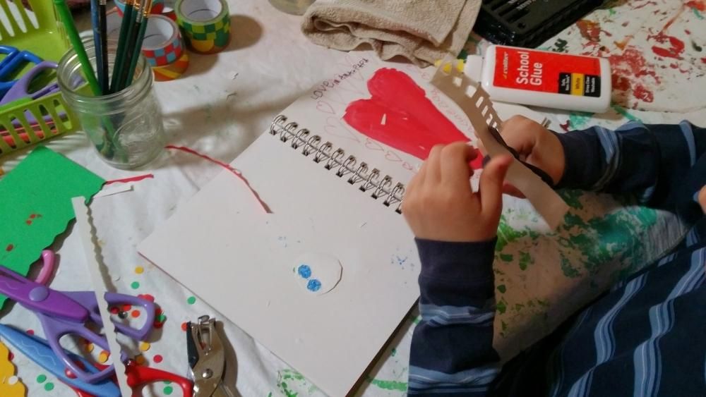 Child sits next to a notebook that they're apparently gluing paper scrap into. The notebook has a heart painted in it with many craft supplies spread around on the table