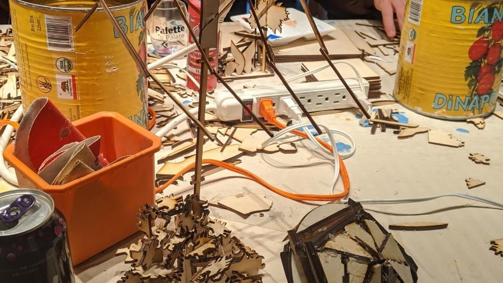 A chaotic selection of wooden bits, power cords, metal cans and paint are spread out on a table