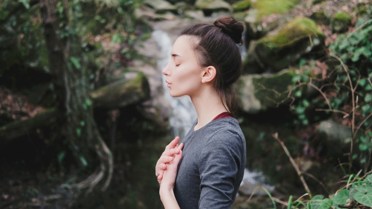 Woman outside near waterfall taking a deep breath with hands over heart.