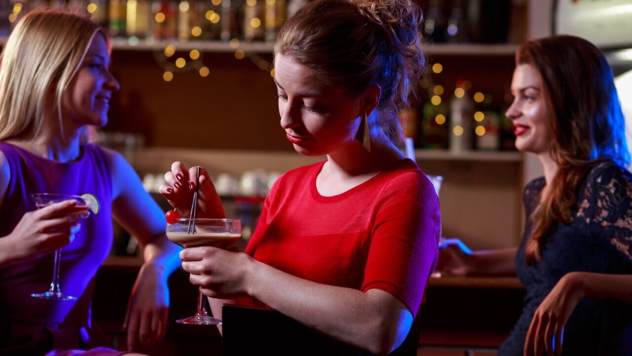 Two friends at bar talking behind another woman who is sad.